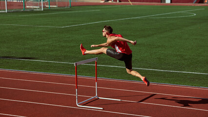 Athlete in red sprinting and jumping over hurdle on green stadium track. Concept of motion, power,...