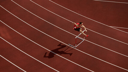 Aerial view of athlete jumping over hurdle on red running track with shadow. Concept of speed, discipline, determination for sports branding, motivation design, and athletic advertising visuals.