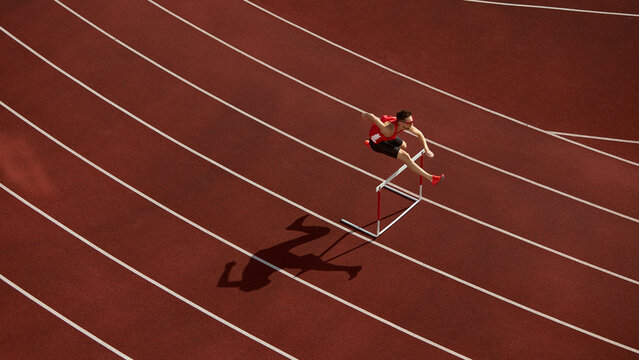 Aerial view of athlete jumping over hurdle on red running track with shadow. Concept of speed, discipline, determination for sports branding, motivation design, and athletic advertising visuals. - Powered by Adobe