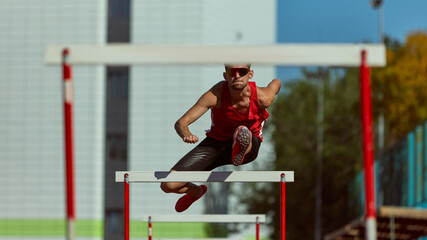 Male hurdler in powerful jump over barrier with city stadium in background. Concept of confidence, progress, physical mastery for advertising, editorial, and sports promotions.