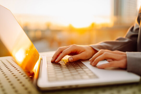 A close-up of a young woman's hands typing on a laptop while sitting at sunset. A female freelancer works on a laptop and admires the sunset from a rooftop terrace. Concept of remote work, relaxation.