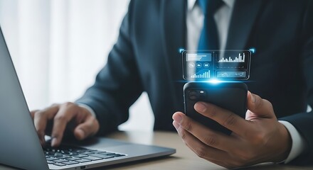 Businessman using smartphone with glowing digital interface next to laptop