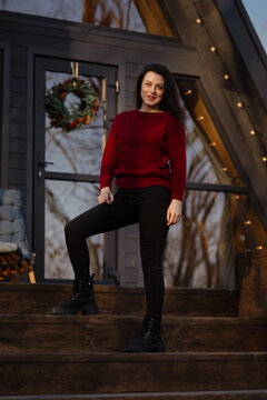 Woman posing confidently on wooden steps of modern home during sunset in cozy winter outfit