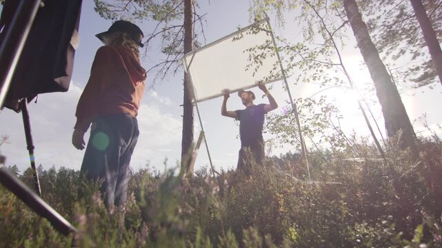 Videographer softens direct sunlight on subject using diffusion sheet in woods