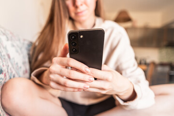 Young Caucasian woman with long brown hair sitting on a couch, focused on her smartphone. She appears engaged and relaxed in a cozy indoor setting.