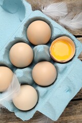 Raw chicken eggs in egg carton and feathers on wooden table, top view