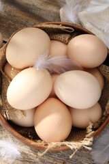 Raw chicken eggs in bowl and feathers on wooden table, top view