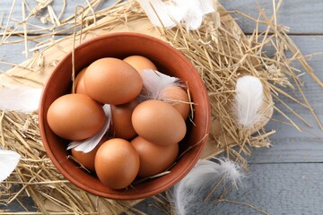 Raw chicken eggs in bowl, feathers and straw on grey wooden table, flat lay. Space for text