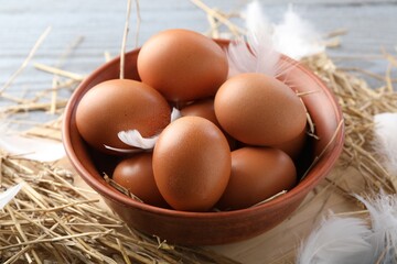 Raw chicken eggs in bowl, feathers and straw on table, closeup