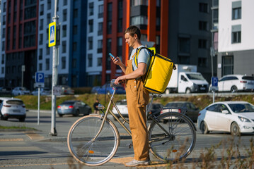 Man standing with bicycle on an urban street, checking his smartphone for delivery information, wearing an insulated yellow backpack for delivering food or packages