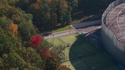 Aerial view of outdoor tennis courts beside a large stadium, surrounded by vibrant autumn forest colors in an urban park setting in Kaunas, Lithuania. - Powered by Adobe