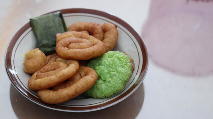 Assorted Indonesian traditional snacks on a plate, featuring fried spiral pastries, pandan rice cake, and banana leaf-wrapped treat. Ideal for culinary visuals and cultural food themes.