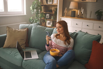 A young woman sits comfortably on a couch, enjoying a bowl of snacks while checking her smartphone....