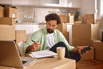 A man sits on the floor of his new home, surrounded by unpacked boxes. He is reviewing paperwork and writing notes while holding a phone and a coffee cup.