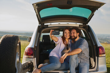 A couple sits in the trunk of their vehicle, smiling and taking a selfie together. The sunny day and open landscape create a joyful outdoor picnic atmosphere.