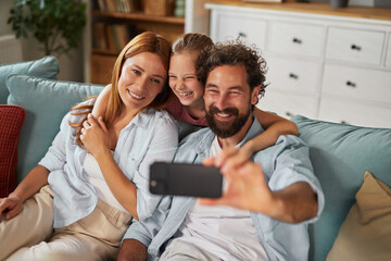Fototapeta premium A joyful family sits together on a comfortable couch, smiling happily as they capture a selfie. The child laughs while the parents embrace her, creating a loving atmosphere.