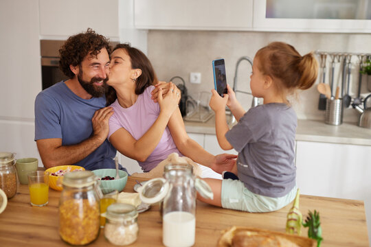 A loving family shares breakfast in the kitchen, filled with laughter and joy. A child takes a picture of her parents while they smile warmly at each other, creating lasting memories.