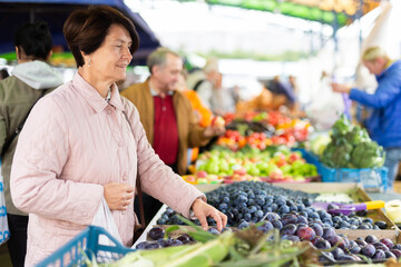 Positive mature woman in casual clothes picking fresh plums during shopping at grocery market