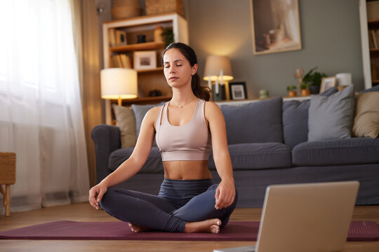 A woman practices meditation at home in a serene living room, seated on a yoga mat with a laptop nearby. Soft lighting enhances the peaceful atmosphere.