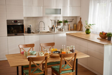 Four chairs are arranged around a wooden table filled with bowls of fruits, cereals, and snacks. The bright kitchen has soft natural light streaming through the window, creating a warm atmosphere.