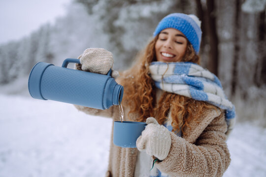 A young woman with a thermos sits on a log in a snowy forest. A cheerful woman enjoys the winter landscape on a sunny day. Concept: drinks, nature, adventure.