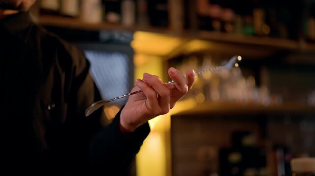 Close-up of a bartender in a dimly lit bar twirling a twisted bar spoon with a muddler end, preparing a craft cocktail amid blurred bottles and glassware on shelves.