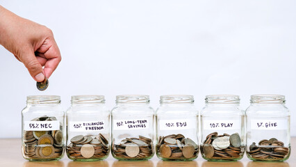 Hand putting coin into one of six labeled glass jars for budgeting plan, representing personal...