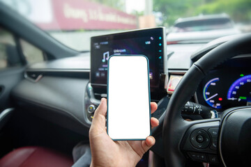 A person holding a smartphone with a blank white screen inside a modern car, focusing on the steering wheel and dashboard. Concept for car navigation app, mobile technology, safe driving awareness.