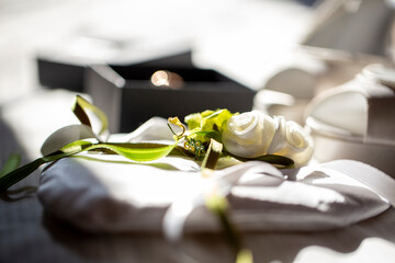 Soft-focus, bright image of a white satin ring bearer pillow with a delicate white rose and green ribbon, next to an open, black jewelry box.