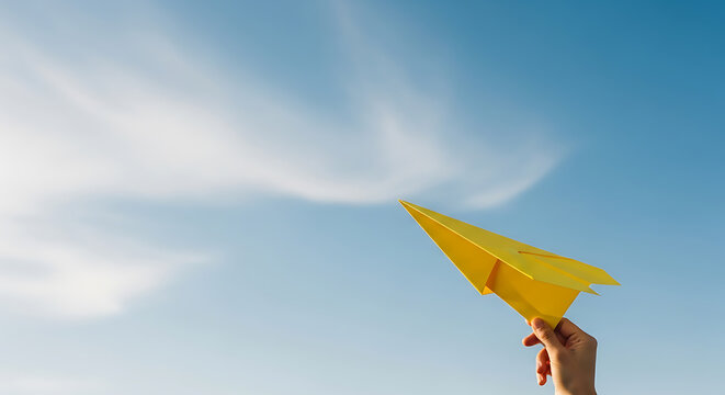 Person holding a yellow paper airplane up to a bright blue sky with light clouds symbolizing dreams and ambition. - Powered by Adobe