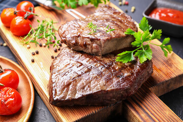 Delicious grilled beef steaks with herbs, peppercorns and tomatoes on dark textured table, closeup