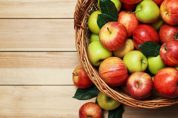 Fresh apples in wicker basket and green leaves on wooden table, top view. Space for text
