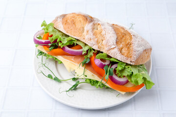 Delicious ciabatta sandwich with prosciutto, cheese and vegetables on white tiled table, closeup