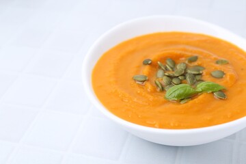 Delicious pumpkin soup with seeds and basil in bowl on white tiled table, closeup. Space for text