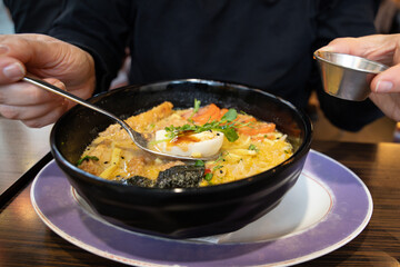 A woman eats ramen, Japanese miso ramen noodles with fried pork and egg, drizzled with sauce.