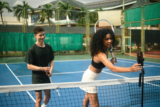 African woman and Asian man tennis players setting up a smartphone on a tripod to record their training session on court