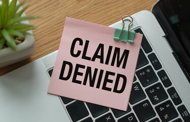 Pink card with the text CLAIM DENIED in a craft envelope on a work desk with a modern laptop keyboard and burgundy pen. Flat layout of the workplace.