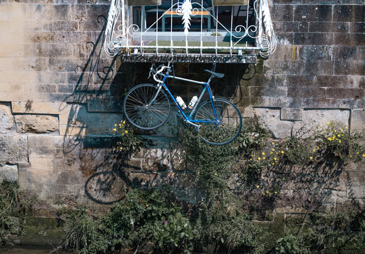 A bicycle hanging below a balcony in an urban setting, likely due to limited space. The scene shows old natural stone walls, giving a rustic and authentic city atmosphere.age
