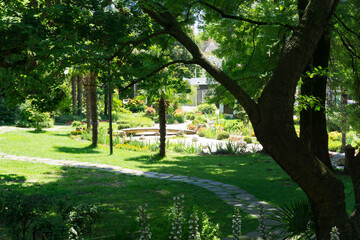 Tranquil garden scene with lush green lawns, tall trees providing shade, and winding stone path leading to vibrant flower bed and small pond. The Russian-Japanese Friendship Garden in Sochi