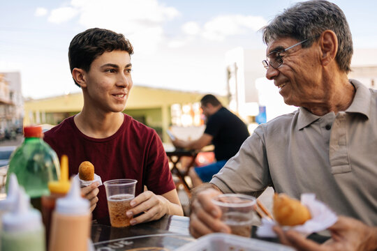 Grandfather and teenage grandson enjoying traditional Brazilian snacks — coxinha and sausage roll — while drinking guarana soda at a street food stall
