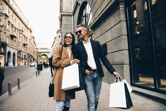 Fashionable couple walking in city with shopping bags, laughing and enjoying time together. Man in black jacket and white shirt,  Asian woman in camel coat and sunglasses. Black friday sales.