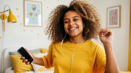 Happy teenage girl with afro hair dancing while listening to music with earphones in a bright bedroom holding a smartphone and enjoying joyfully. - Powered by Adobe