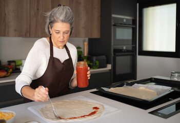 Woman spreading tomato sauce on pizza dough for a homemade meal in kitchen
