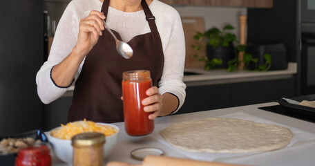 Woman in apron spreading tomato sauce on pizza dough in a modern kitchen