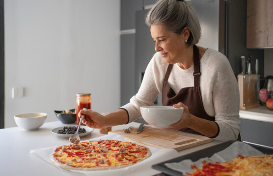 Senior woman preparing pizza, adding toppings on dough in a kitchen