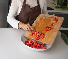 Woman in apron chopping red tomato on wooden board, adding to a bowl for salad