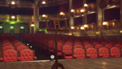 Under soft lights, a vacant theater stage is ready for action. The rich red seats create a warm atmosphere, while the wooden music stand stands as the only sign of preparation for upcoming shows.