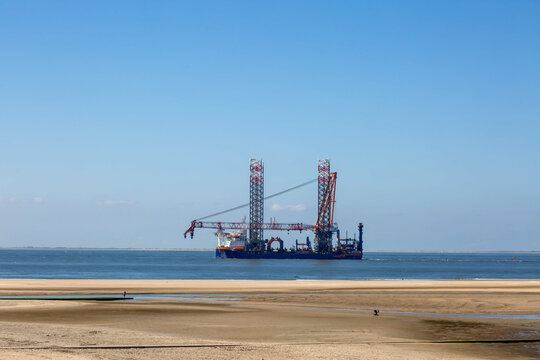 Ein grosses Offshore Schiff mit hohen Tuermen und Kran liegt nahe dem Strand unter blauem Himmel auf der Nordseeinsel Borkum.