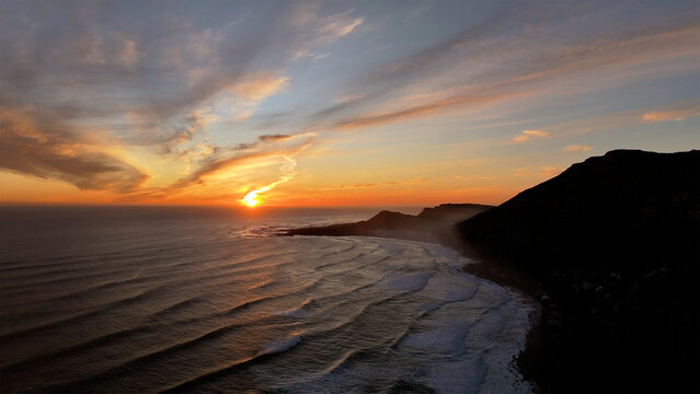 Aerial view of the sun setting in shades of gold, painting the ocean and sandy beach with an ethereal glow beneath the dark silhouette of the mountain, Cape Town, Western Cape, South Africa.