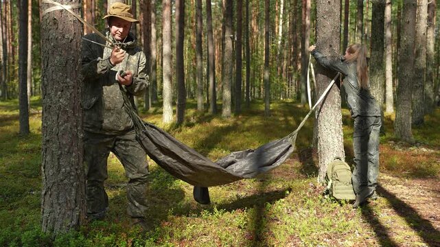 A hammock is being hung between trees by a man and a woman. In the forest, a hammock is being set up. In the woods, a hiking couple is relaxing with a hammock. Travelers wearing camouflage clothing